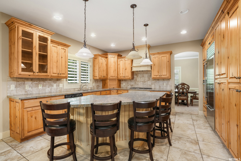 Kitchen featuring appliances with stainless steel finishes, a center island, decorative light fixtures, light stone counters, custom exhaust hood, brown cabinets, a center island with sink, backsplash, and light tile floors