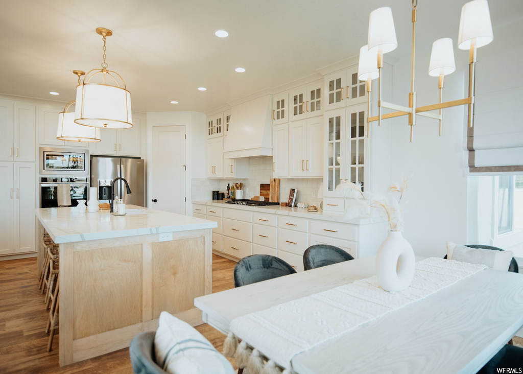 Kitchen featuring white cabinets, custom exhaust hood, hardwood flooring, backsplash, hanging light fixtures, appliances with stainless steel finishes, and light countertops