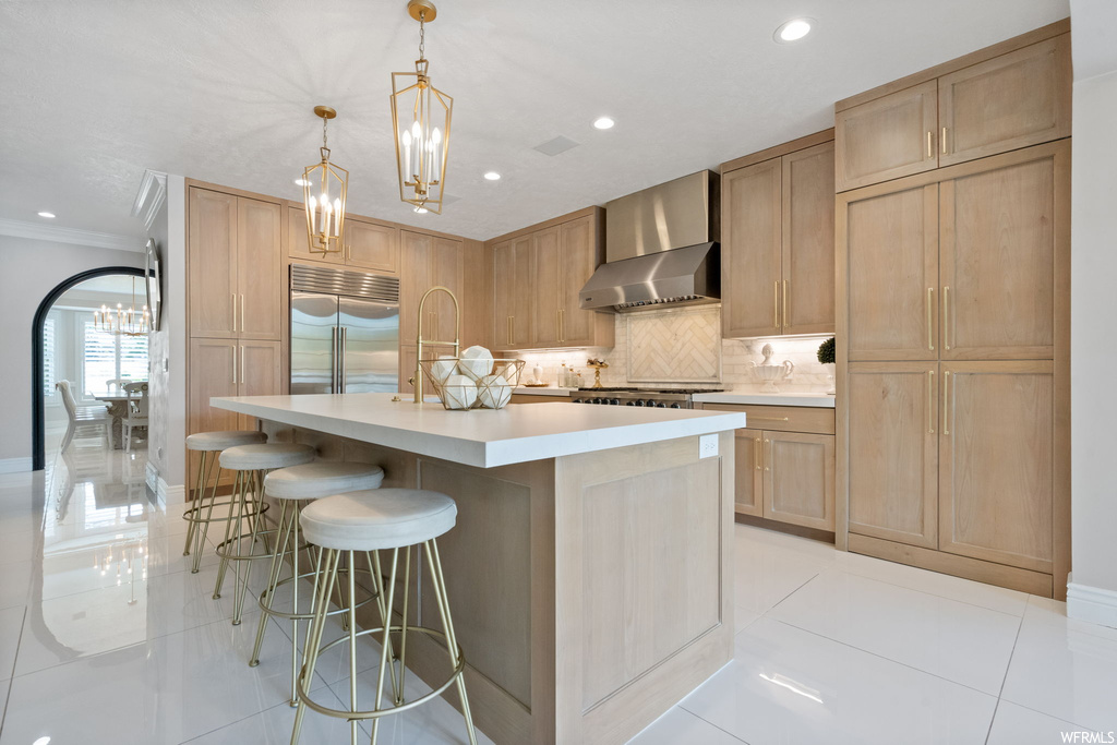 Kitchen featuring light tile floors, stainless steel appliances, backsplash, ornamental molding, light countertops, decorative light fixtures, kitchen island with sink, wall chimney exhaust hood, a kitchen island, and light brown cabinets