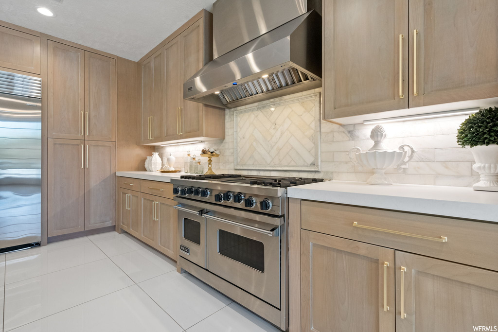 Kitchen with light tile flooring, premium appliances, wall chimney exhaust hood, light countertops, and backsplash