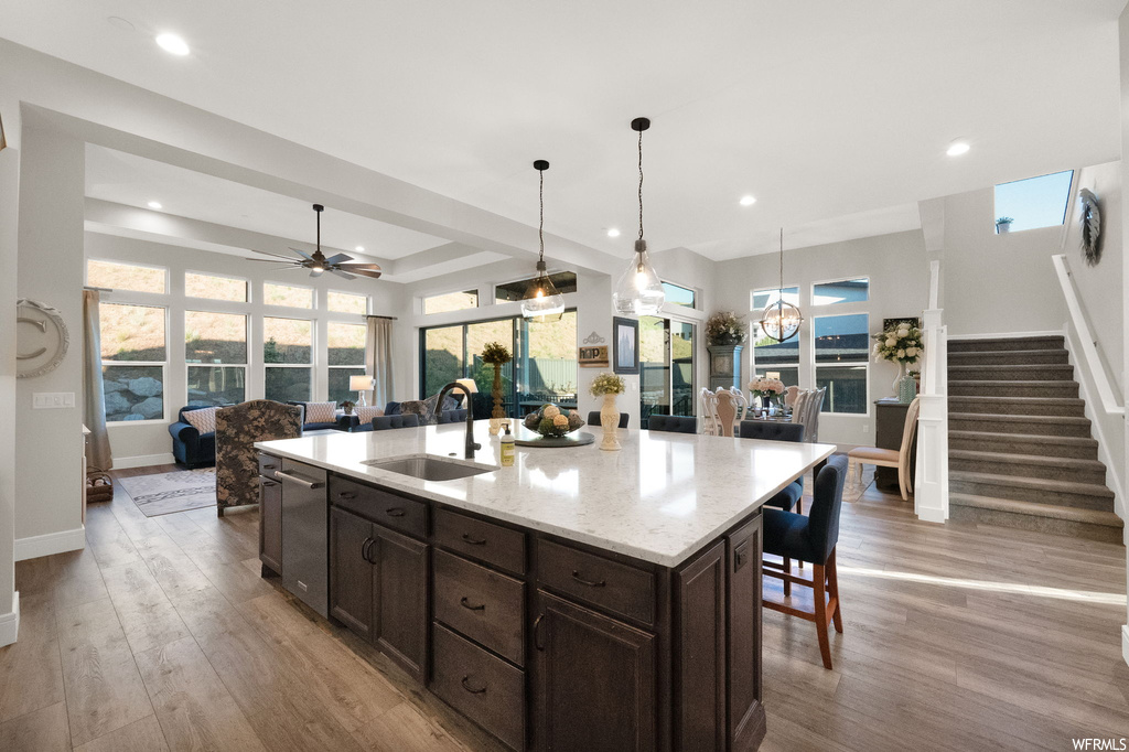 Kitchen with dark brown cabinets, ceiling fan, light hardwood floors, pendant lighting, a tray ceiling, kitchen island with sink, and stainless steel dishwasher