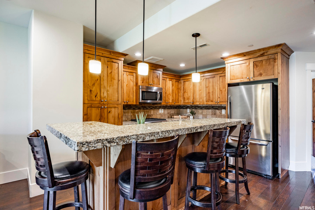 Kitchen featuring light stone counters, brown cabinets, pendant lighting, backsplash, a center island, appliances with stainless steel finishes, and dark hardwood flooring