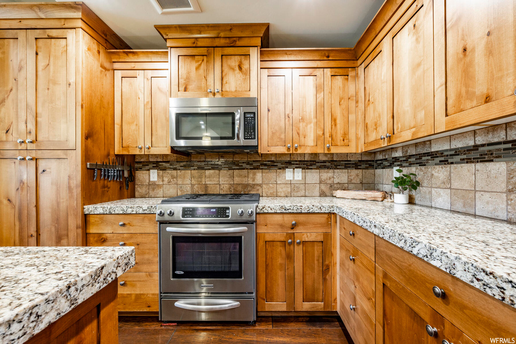 Kitchen featuring stainless steel appliances, brown cabinets, backsplash, light granite-like countertops, and dark hardwood flooring