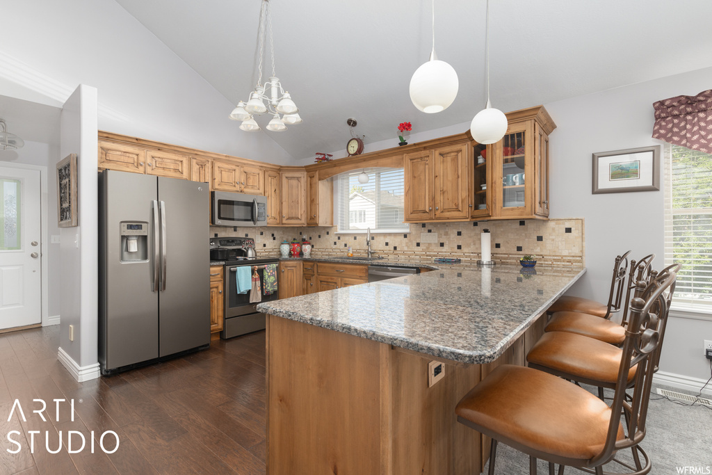 Kitchen featuring stainless steel appliances, brown cabinets, granite-like countertops, hanging light fixtures, backsplash, vaulted ceiling, hardwood flooring, a center island, and plenty of natural light