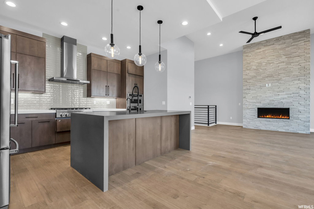 Kitchen featuring wall chimney range hood, light hardwood / wood-style flooring, pendant lighting, ceiling fan, and a fireplace