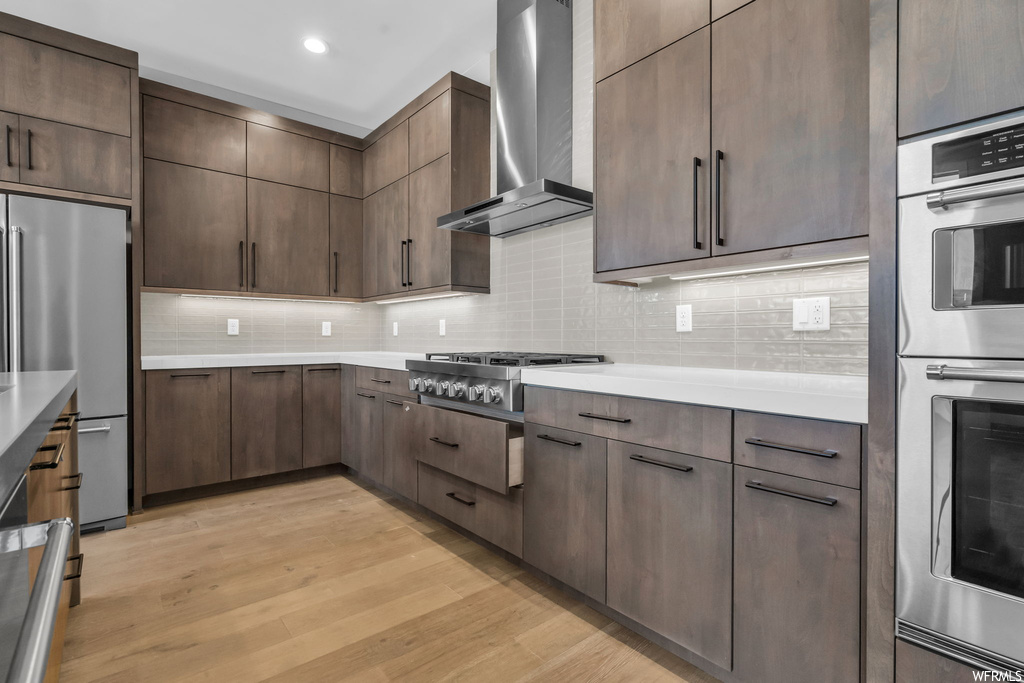Kitchen with light hardwood / wood-style flooring, stainless steel appliances, wall chimney exhaust hood, tasteful backsplash, and dark brown cabinetry