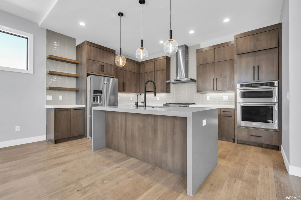 Kitchen featuring wall chimney exhaust hood, a center island with sink, light wood-type flooring, and backsplash