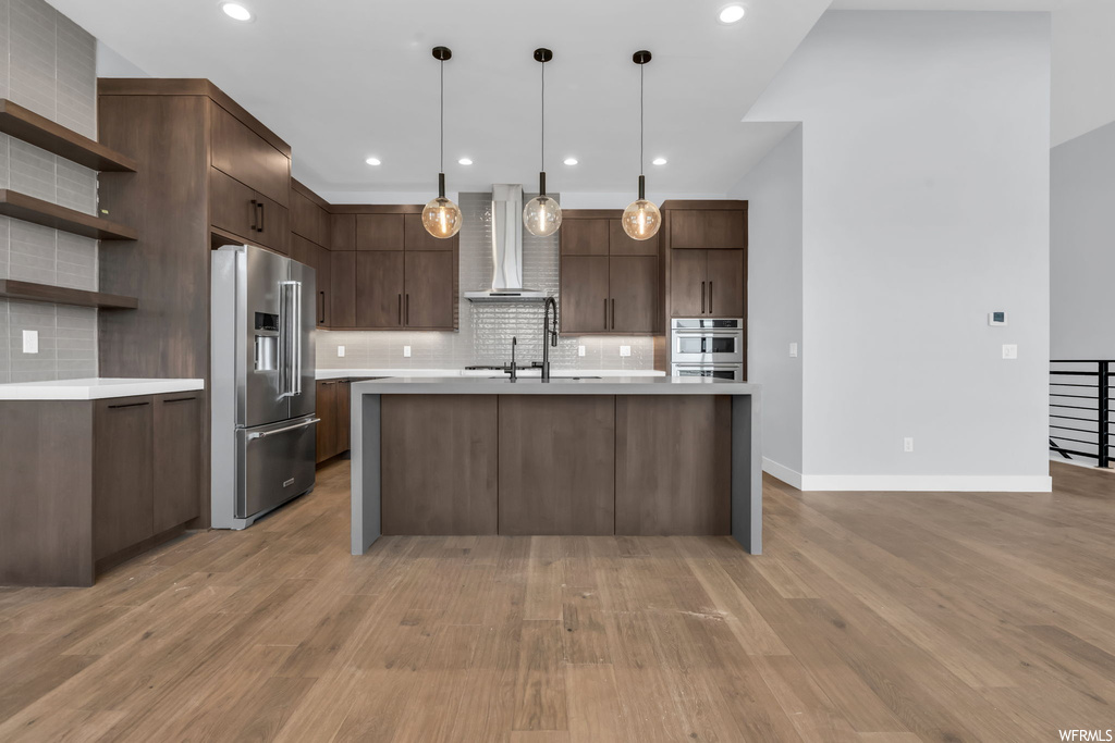 Kitchen with wall chimney exhaust hood, pendant lighting, stainless steel appliances, backsplash, and light wood-type flooring