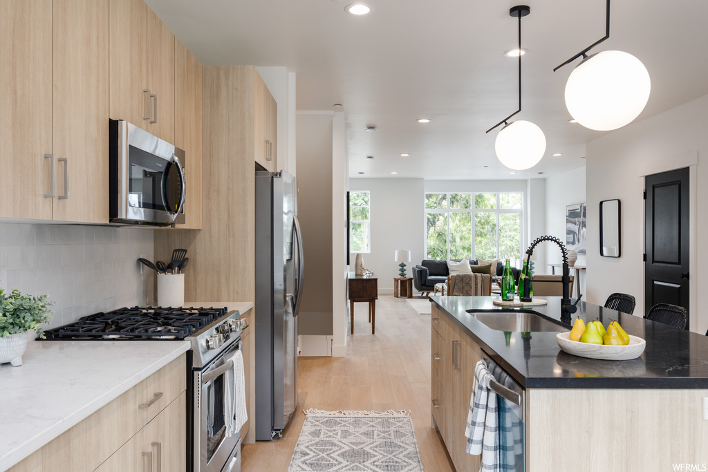 Kitchen with light hardwood flooring, stainless steel appliances, pendant lighting, dark countertops, and backsplash