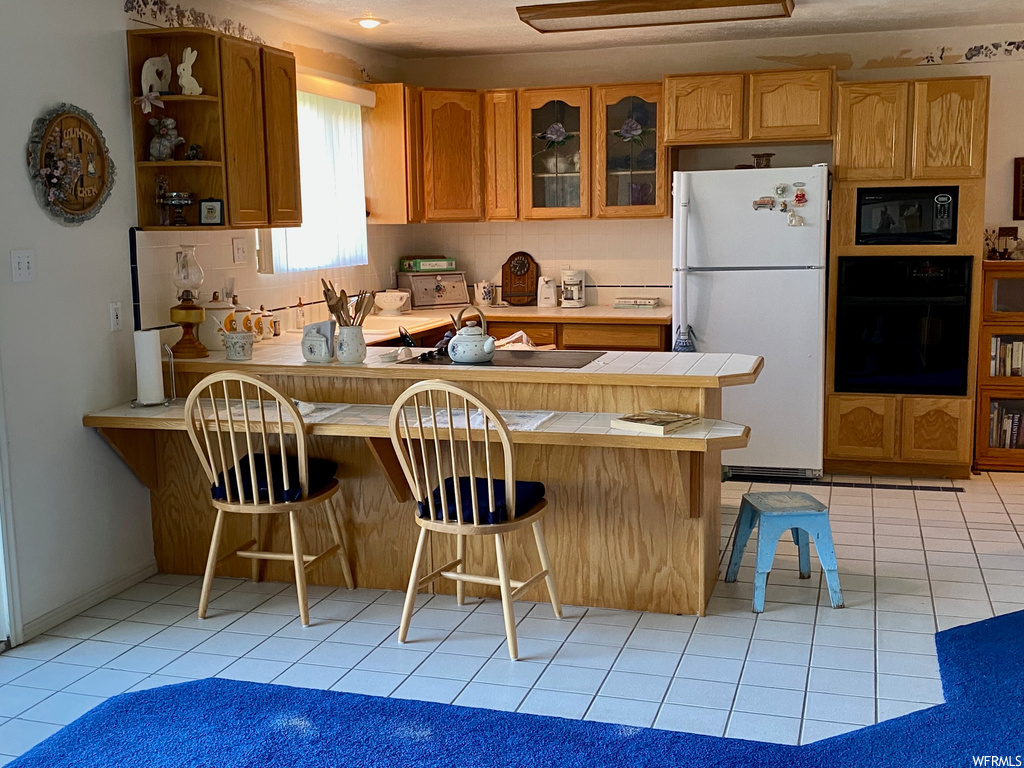 Kitchen with light tile floors, kitchen peninsula, white fridge, and black microwave