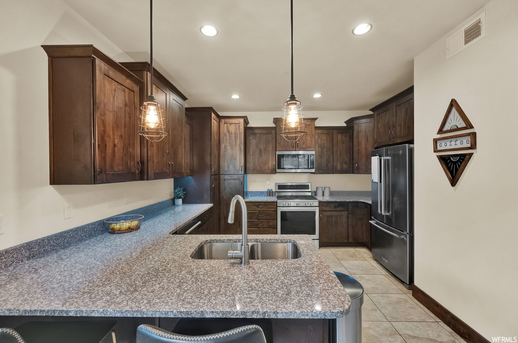 Kitchen featuring light tile flooring, dark brown cabinetry, hanging light fixtures, stone counters, and appliances with stainless steel finishes