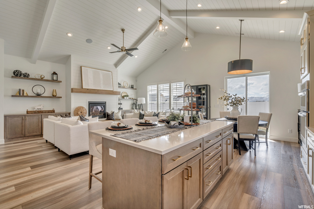 Kitchen featuring plenty of natural light, light hardwood flooring, pendant lighting, a center island, a high ceiling, ceiling fan, light countertops, a fireplace, and vaulted ceiling with beams