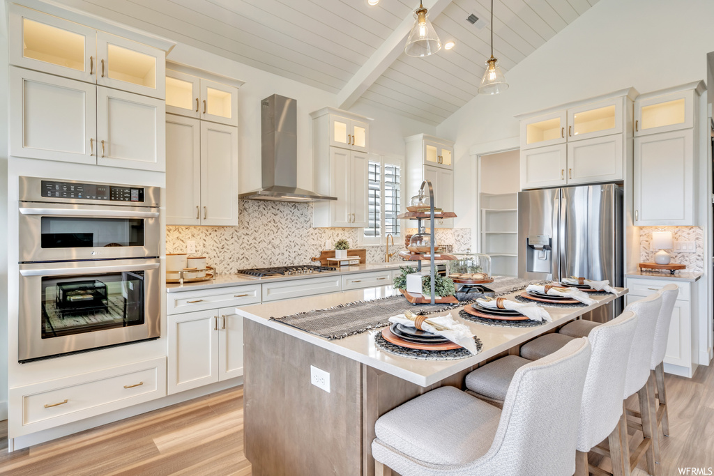 Kitchen featuring light hardwood floors, a center island with sink, a high ceiling, pendant lighting, backsplash, wall chimney exhaust hood, vaulted ceiling with beams, appliances with stainless steel finishes, a kitchen island, and white cabinetry