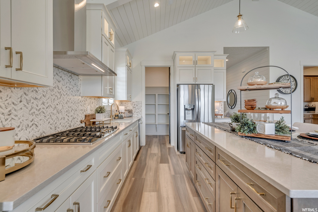 Kitchen featuring wall chimney range hood, vaulted ceiling, hanging light fixtures, stainless steel gas stovetop, a high ceiling, backsplash, light countertops, light parquet floors, and white cabinets