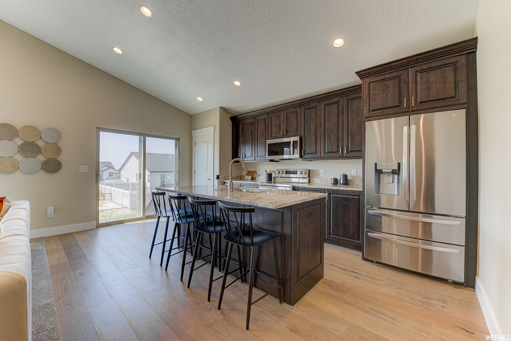Kitchen with appliances with stainless steel finishes, a center island, light stone counters, vaulted ceiling, dark brown cabinets, and light hardwood flooring