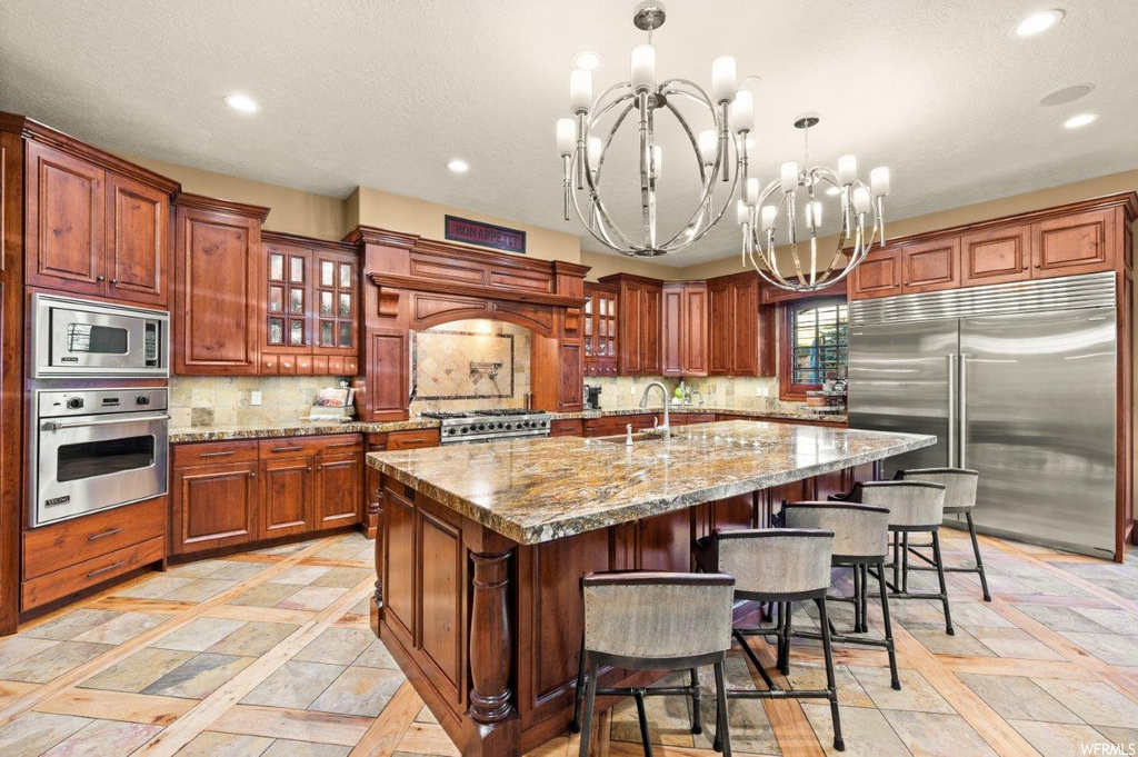 Kitchen with a chandelier, a center island with sink, a center island, backsplash, light tile floors, brown cabinets, stone counters, and built in appliances