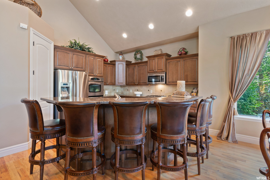 Kitchen with brown cabinets, light granite-like countertops, lofted ceiling, appliances with stainless steel finishes, backsplash, and light parquet floors