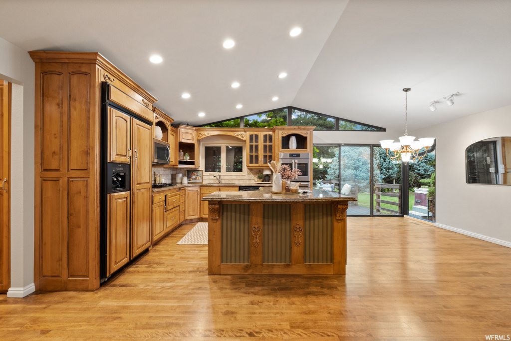 Kitchen featuring stainless steel oven, an island with sink, paneled built in fridge, brown cabinets, vaulted ceiling, light hardwood flooring, and backsplash