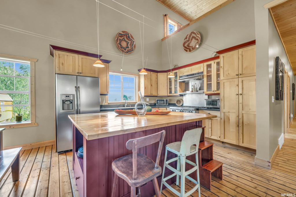 Kitchen with a center island, stainless steel refrigerator with ice dispenser, wooden ceiling, a wealth of natural light, vaulted ceiling, light countertops, light hardwood flooring, and a high ceiling