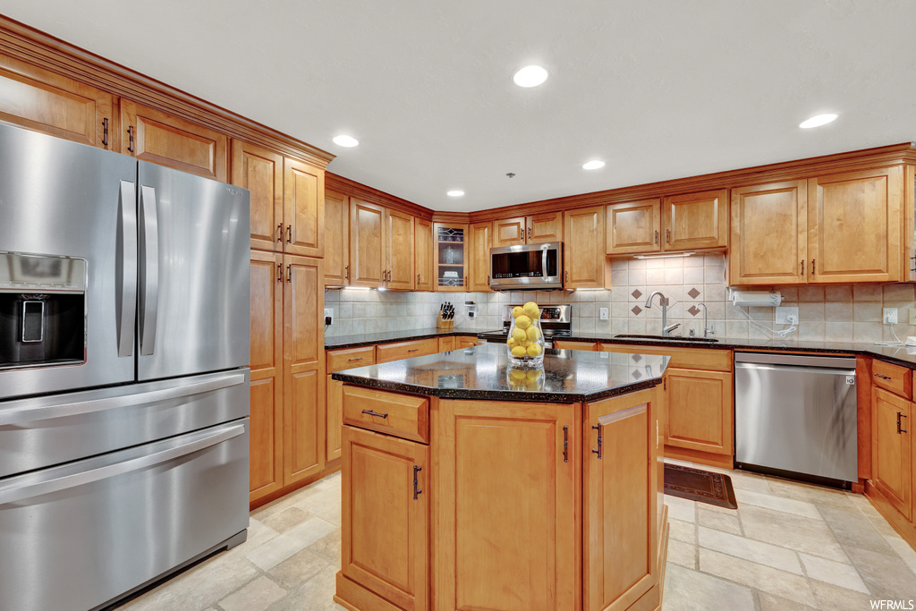 Kitchen with dark countertops, backsplash, light tile floors, brown cabinets, appliances with stainless steel finishes, and a kitchen island with sink