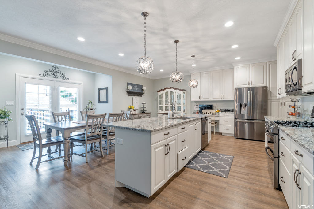 Kitchen with light granite-like countertops, crown molding, pendant lighting, an island with sink, white cabinetry, stainless steel appliances, and light parquet floors