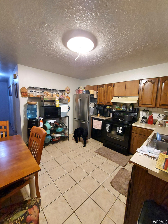 Kitchen with a textured ceiling, light tile flooring, black / electric stove, and stainless steel refrigerator