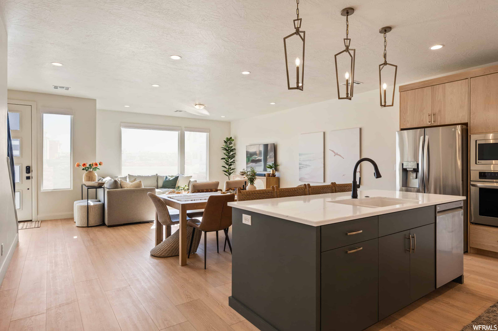 Kitchen featuring appliances with stainless steel finishes, light countertops, hanging light fixtures, light hardwood flooring, and kitchen island with sink