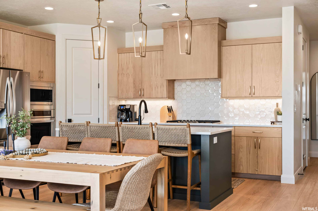 Kitchen featuring stainless steel appliances, a center island, a kitchen island with sink, pendant lighting, light countertops, light hardwood flooring, and backsplash