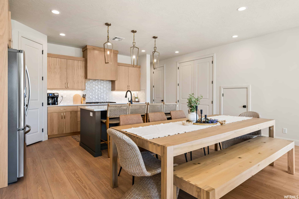 Kitchen featuring a center island, stainless steel fridge, pendant lighting, light countertops, kitchen island with sink, backsplash, and light hardwood floors