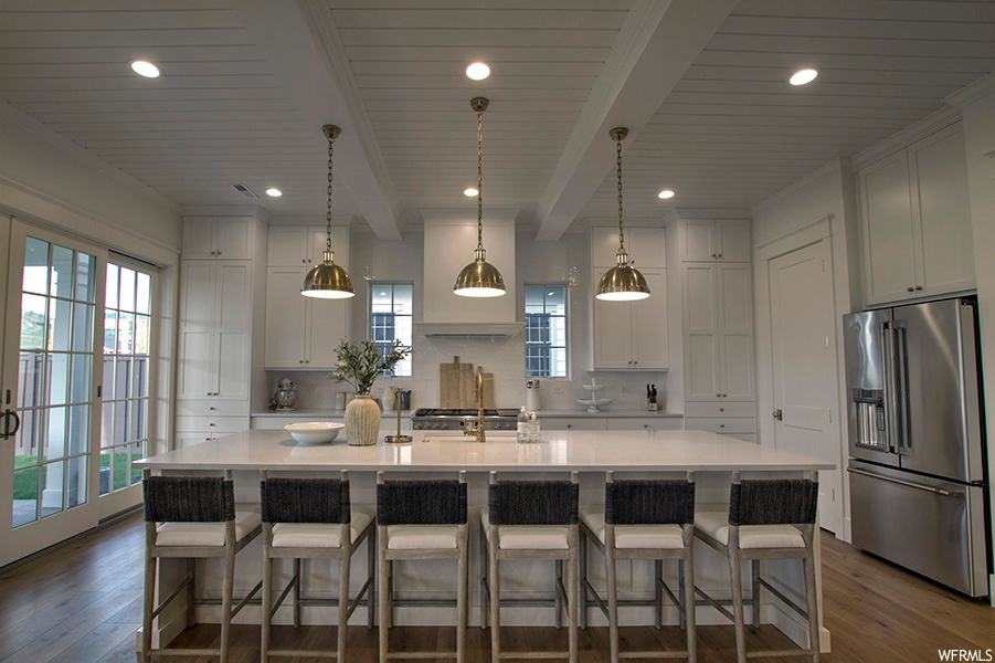 Kitchen with high end fridge, a kitchen island, pendant lighting, white cabinets, dark hardwood flooring, light countertops, backsplash, and beam ceiling