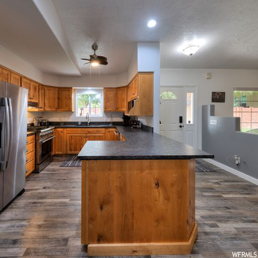 Kitchen with appliances with stainless steel finishes, an island with sink, dark countertops, hardwood floors, brown cabinets, and ceiling fan