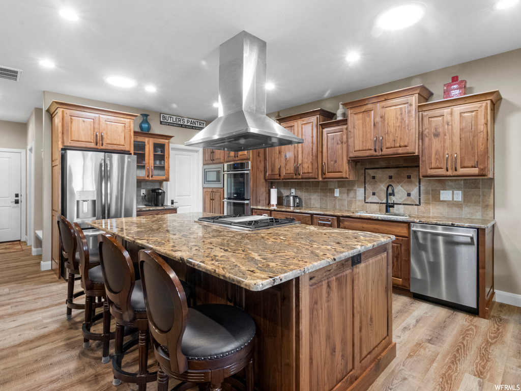 Kitchen featuring appliances with stainless steel finishes, a kitchen island, an island with sink, light stone counters, backsplash, island exhaust hood, and light hardwood floors