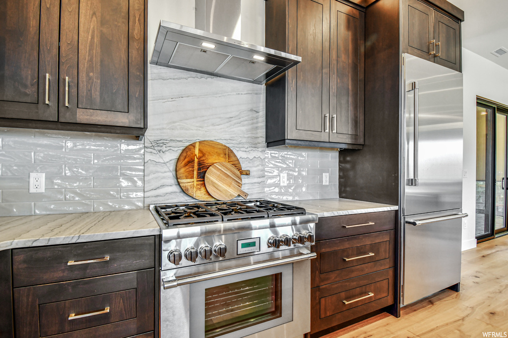 Kitchen featuring wall chimney range hood, tasteful backsplash, high end appliances, light hardwood floors, and dark brown cabinetry