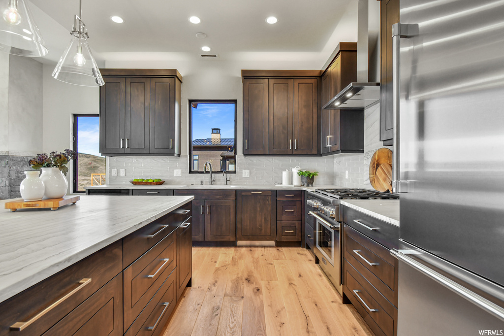 Kitchen featuring wall chimney range hood, light hardwood floors, decorative light fixtures, and backsplash