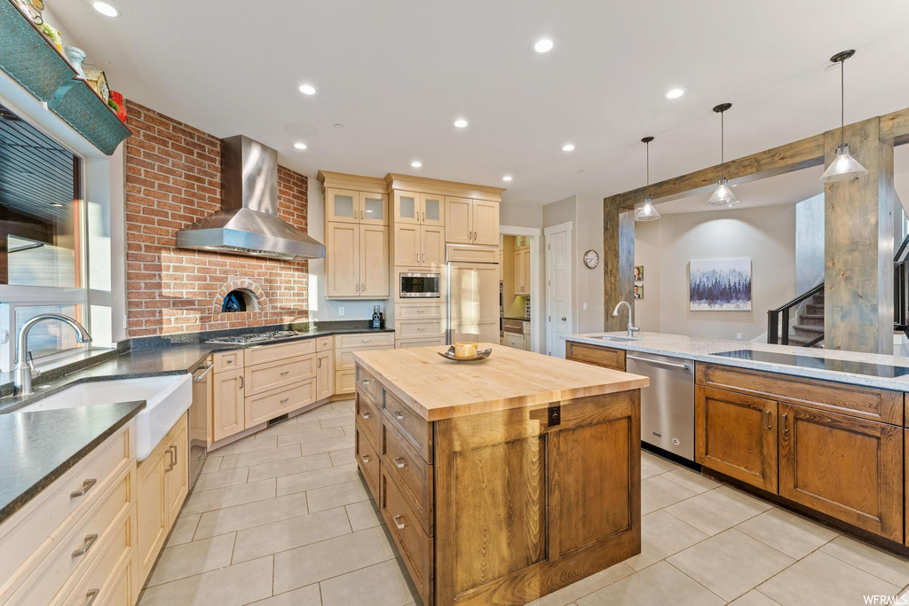 Kitchen featuring brick wall, a kitchen island with sink, wall chimney exhaust hood, built in appliances, light countertops, hanging light fixtures, and light tile floors