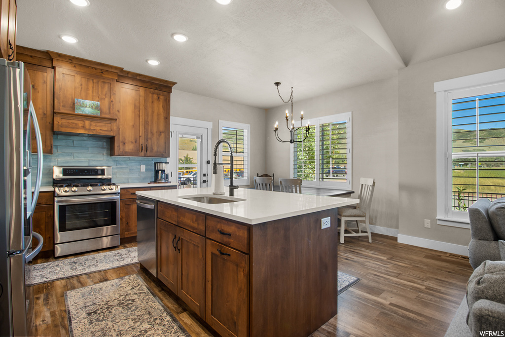 Kitchen with stainless steel appliances, a kitchen island with sink, wood-type flooring, light countertops, and backsplash