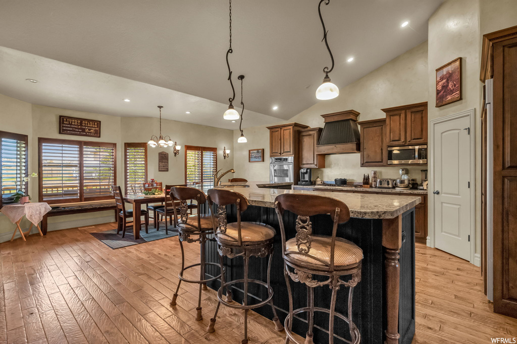 Kitchen featuring appliances with stainless steel finishes, a kitchen island, custom range hood, pendant lighting, lofted ceiling, light stone counters, light hardwood flooring, and a high ceiling