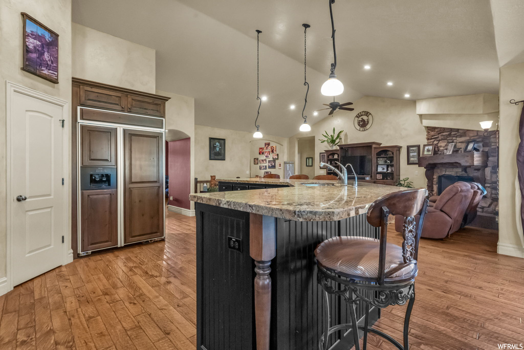 Kitchen with decorative light fixtures, kitchen island with sink, paneled built in fridge, a center island, light hardwood flooring, ceiling fan, light stone countertops, lofted ceiling, a fireplace, and a high ceiling