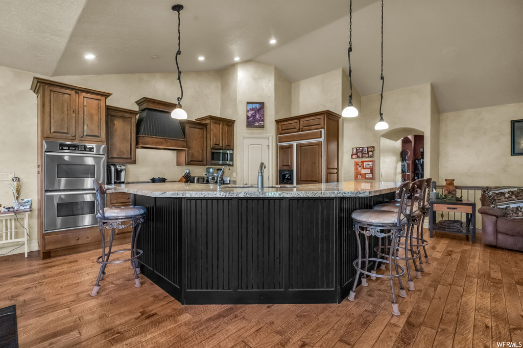 Kitchen featuring hanging light fixtures, vaulted ceiling, light hardwood floors, a center island, premium range hood, dark brown cabinetry, light stone countertops, stainless steel appliances, and a high ceiling