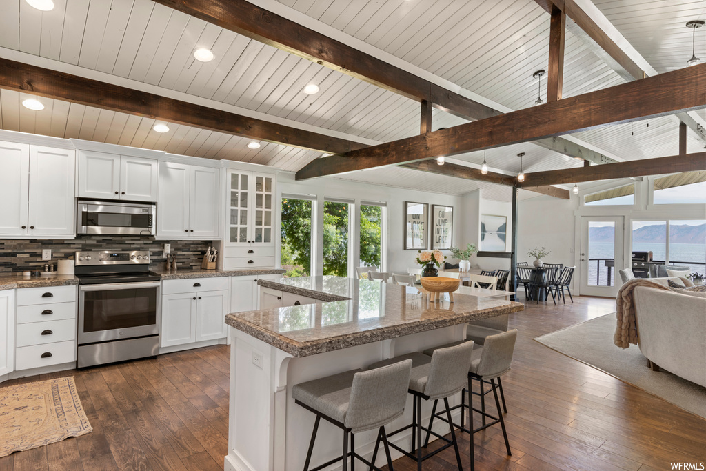 Kitchen featuring backsplash, white cabinets, a kitchen island, hardwood flooring, lofted ceiling with beams, stainless steel appliances, and dark stone counters