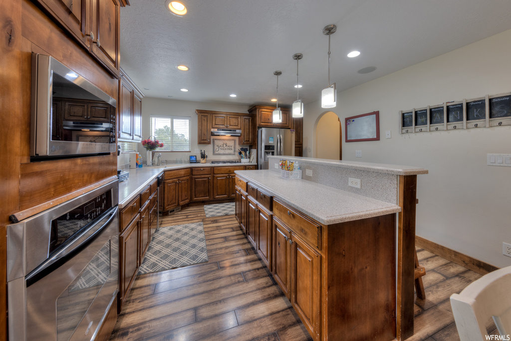 Kitchen with brown cabinets, light countertops, hardwood floors, pendant lighting, and stainless steel appliances