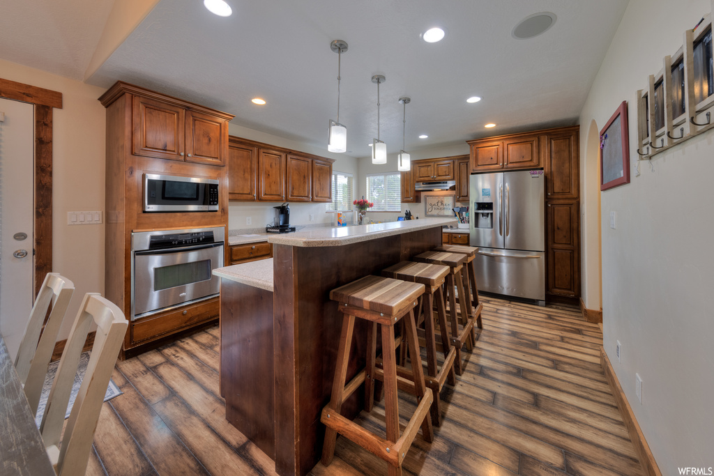 Kitchen with decorative light fixtures, brown cabinets, light countertops, a center island, dark hardwood flooring, and stainless steel appliances