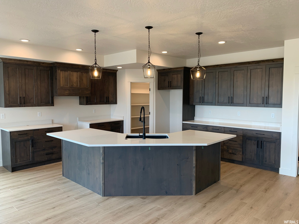 Kitchen featuring dark brown cabinets, light countertops, light hardwood floors, pendant lighting, and a kitchen island with sink