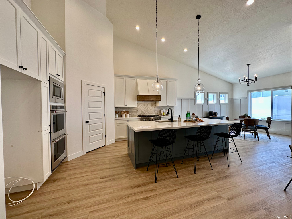Kitchen with custom range hood, a center island, decorative light fixtures, white cabinetry, stainless steel appliances, lofted ceiling, light countertops, a center island with sink, backsplash, light hardwood floors, and a high ceiling