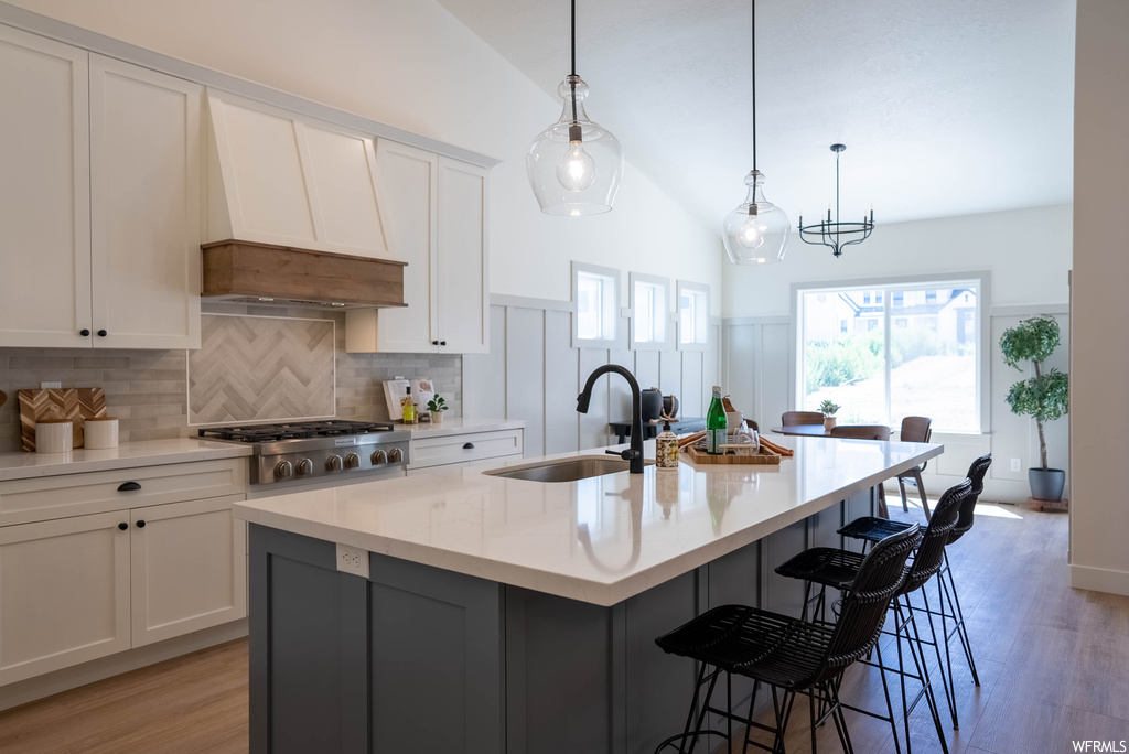 Kitchen with decorative light fixtures, kitchen island with sink, backsplash, light countertops, light hardwood floors, custom exhaust hood, white cabinetry, a center island, lofted ceiling, and stainless steel gas stovetop