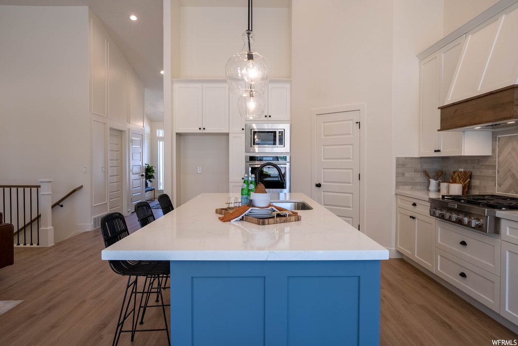 Kitchen with a kitchen island, premium range hood, white cabinetry, decorative light fixtures, stainless steel appliances, wood-type flooring, light countertops, a center island with sink, backsplash, and a high ceiling