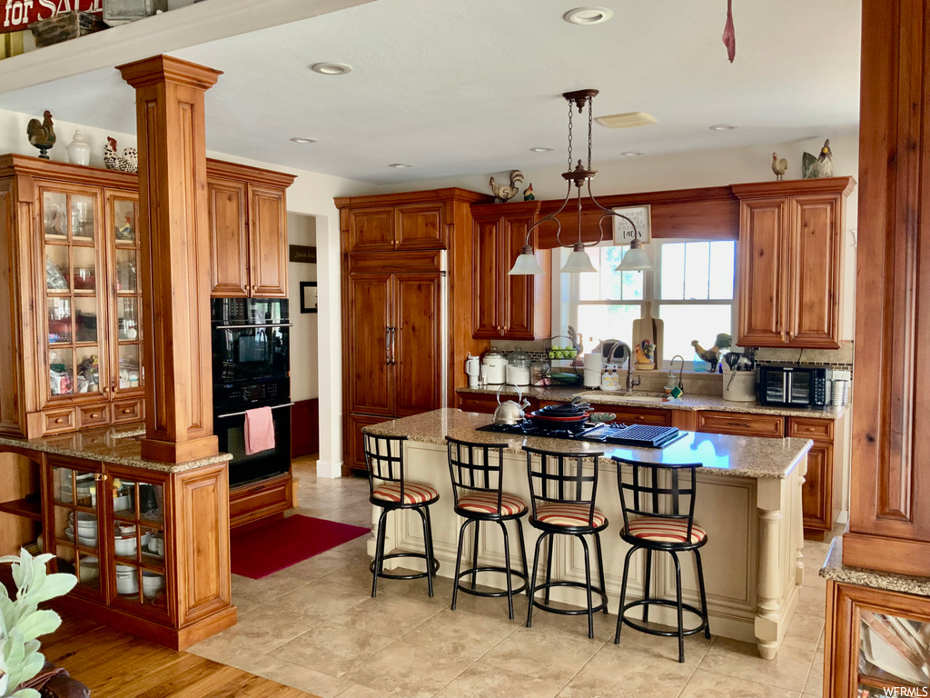 Kitchen featuring gas cooktop, hanging light fixtures, black double oven, backsplash, brown cabinets, a kitchen island, paneled built in refrigerator, light tile floors, and light stone counters