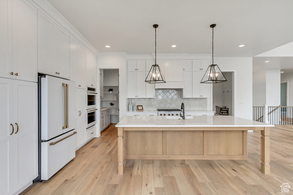 Kitchen featuring white cabinetry, high end fridge, decorative light fixtures, an island with sink, and light wood-type flooring