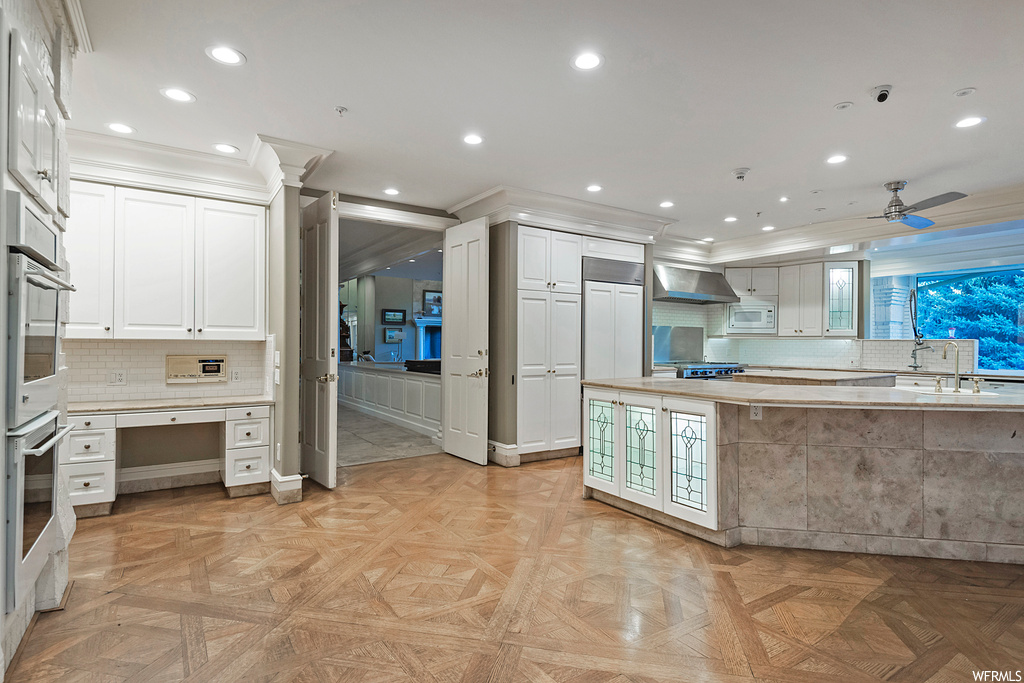Kitchen with exhaust hood, white cabinets, built in appliances, light parquet flooring, light countertops, and backsplash