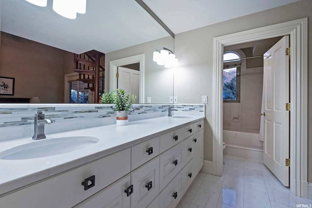Bathroom with backsplash, mirror, light tile floors, and dual bowl vanity